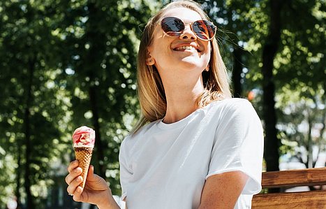 Frau mit Eis in der Hand sitzt auf einer Parkbank in der Sonne.