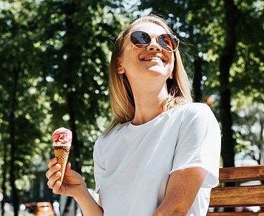 Frau mit Eis in der Hand sitzt auf einer Parkbank in der Sonne.