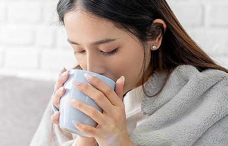 Frau sitzt mit Decke auf dem Sofa und trinkt aus einer Tasse