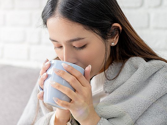 Frau sitzt mit Decke auf dem Sofa und trinkt aus einer Tasse