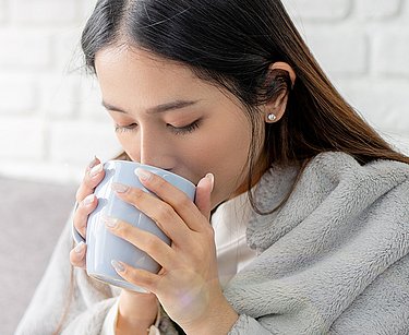 Frau sitzt mit Decke auf dem Sofa und trinkt aus einer Tasse