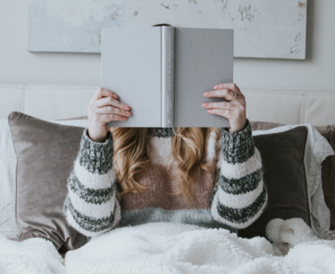 Frau sitzt im Bett mit einem Buch vor dem Gesicht